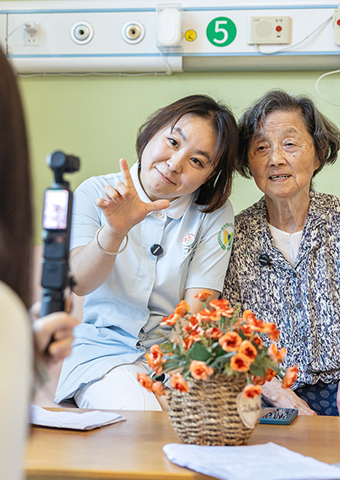 ◎ 初秋,浙江省湖州市蜀山養護院內,幾位頭髮花白的老人正在鏡頭下拍攝系列微短劇《蜀山情報站》。圖為沈婉婷(左)在和一位老人錄製短視頻作品《湖州話等級考試》。(新華社)